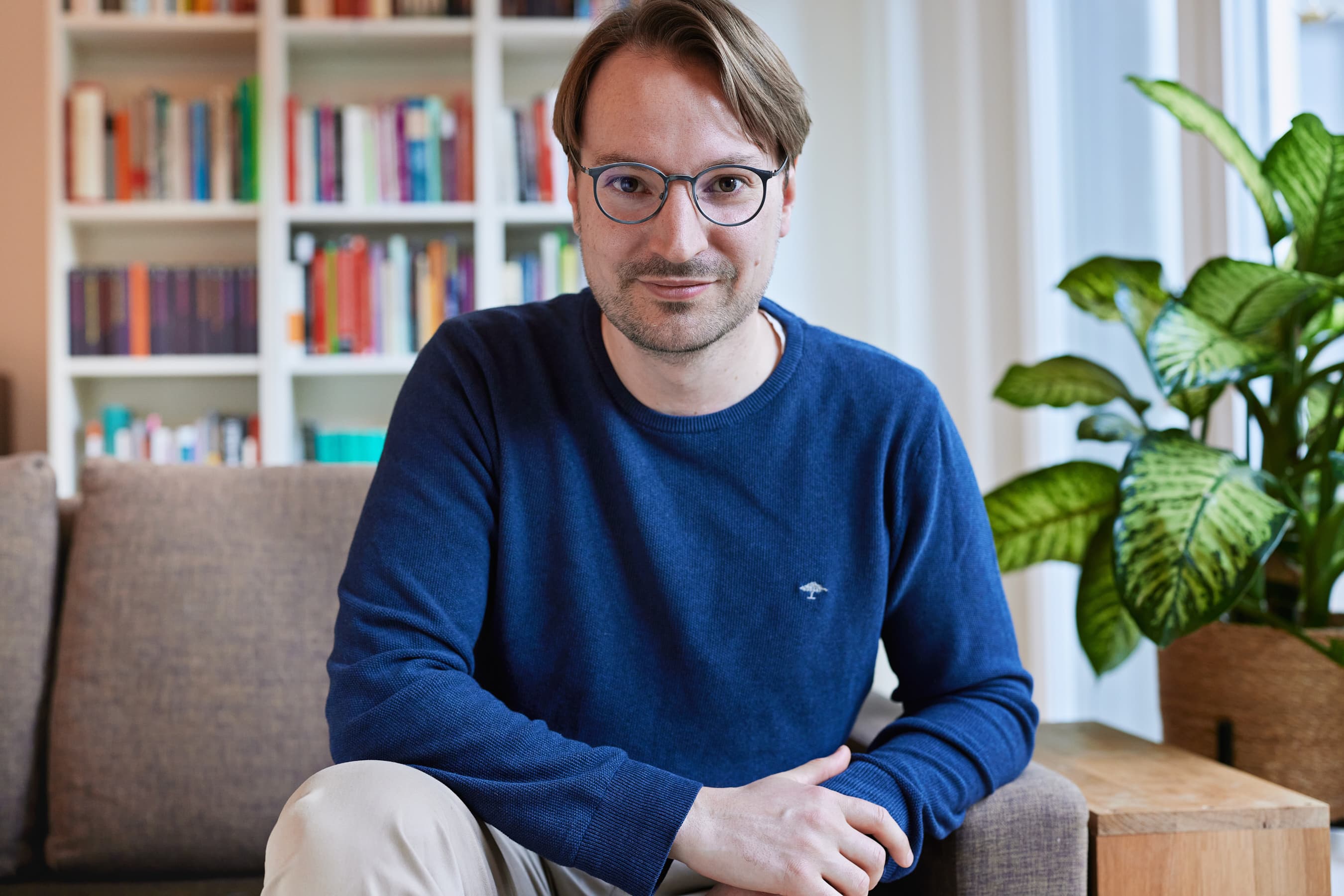 Emanuel Rechsteiner-Grammenos sitzt vorgebeugt auf einem hellen Sofa, die Unterarme auf den Knien, und blickt mit leichtem Lächeln in die Kamera. Er trägt eine runde Brille und einen dunkelblauen Pullover; hinter ihm ein weißes Regal voller bunter Bücher, rechts eine große Grünpflanze.