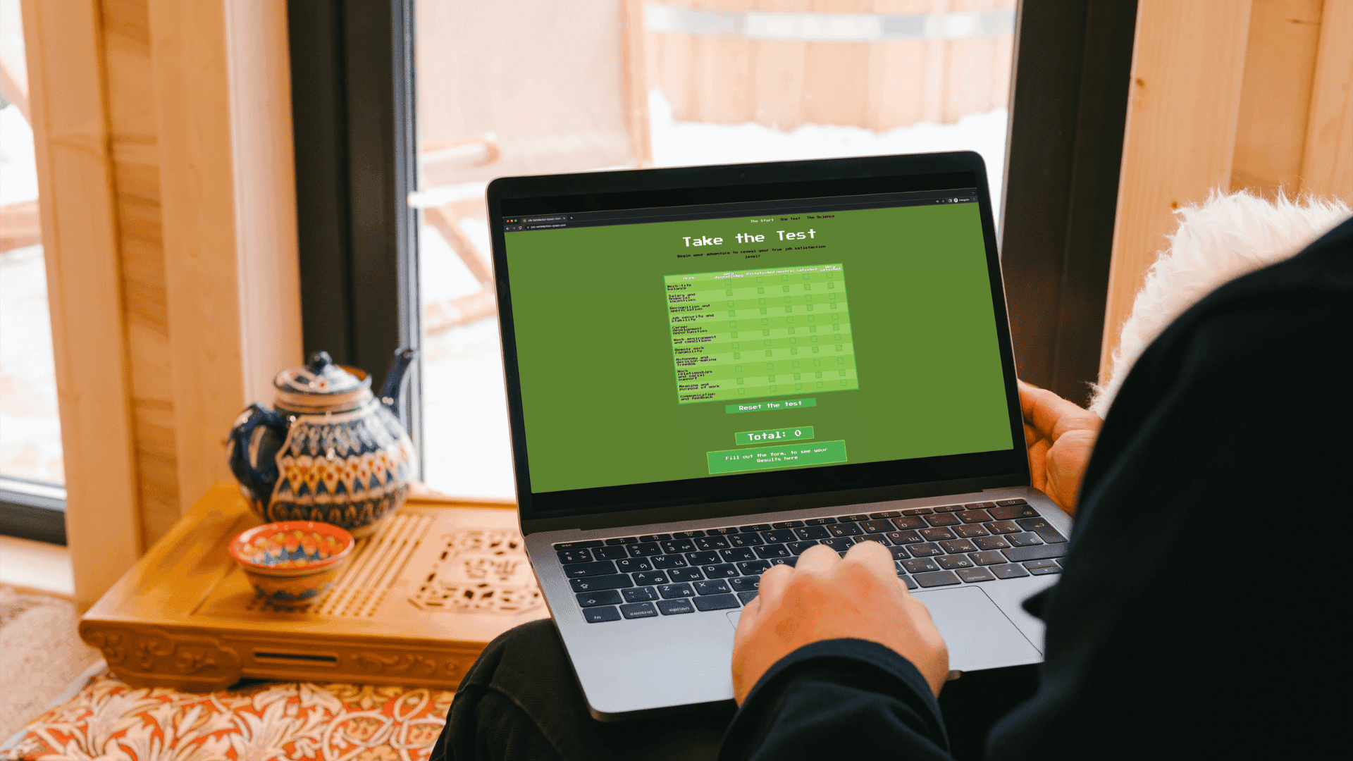 Ein aufgeklappter Laptop auf den Knien einer Person zeigt einen grünen Onlinetest („Take the Test“) mit Fragen und Antwortspalten; daneben auf einem niedrigen Holztisch eine bemalte Teekanne und eine Schale, im Hintergrund ein helles Holzfenster.
