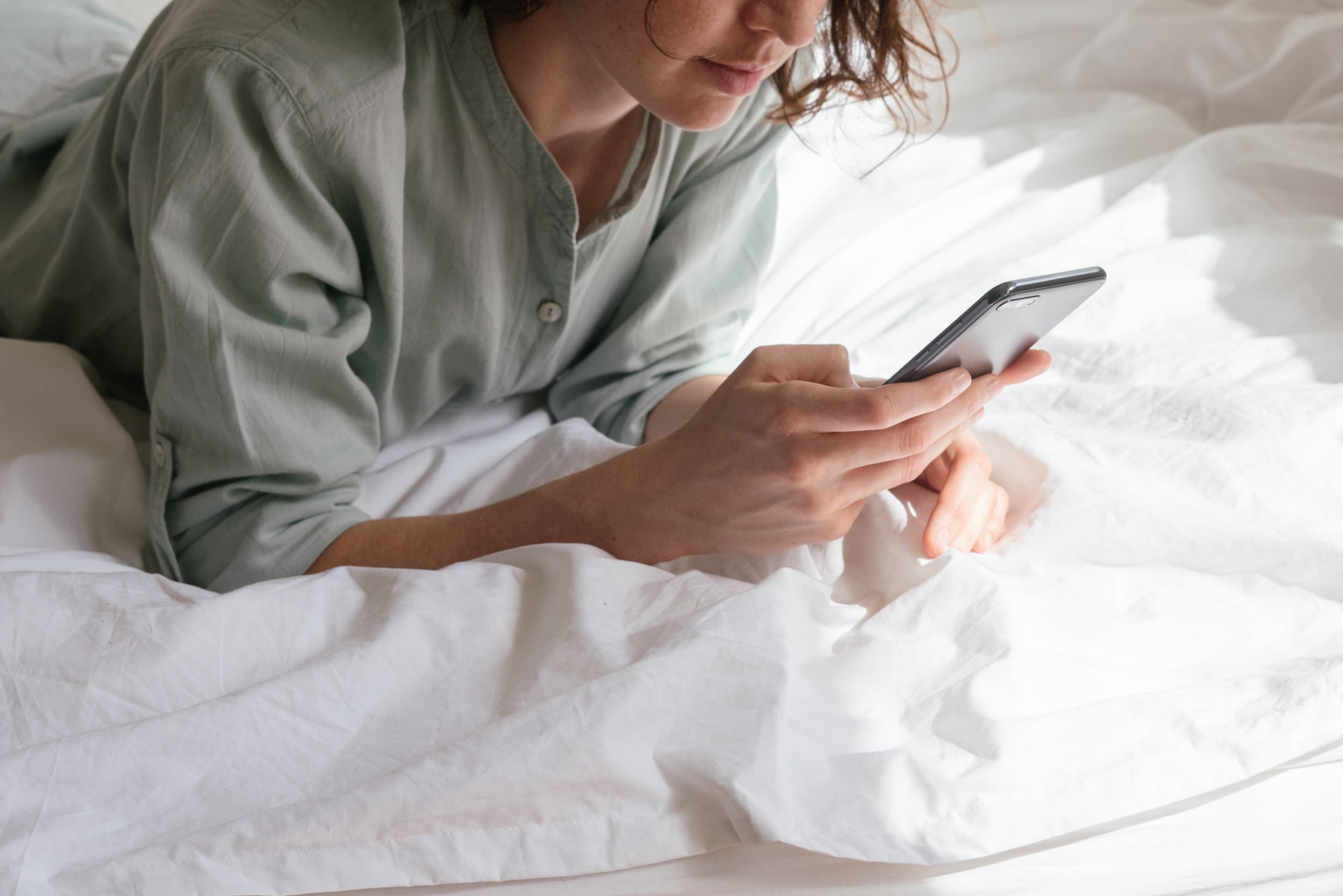 Eine Frau in einem hellgrünen Hemd liegt bäuchlings in weißer Bettwäsche und tippt auf ihrem Smartphone; ihr Gesicht ist nur bis zur Nase zu sehen.