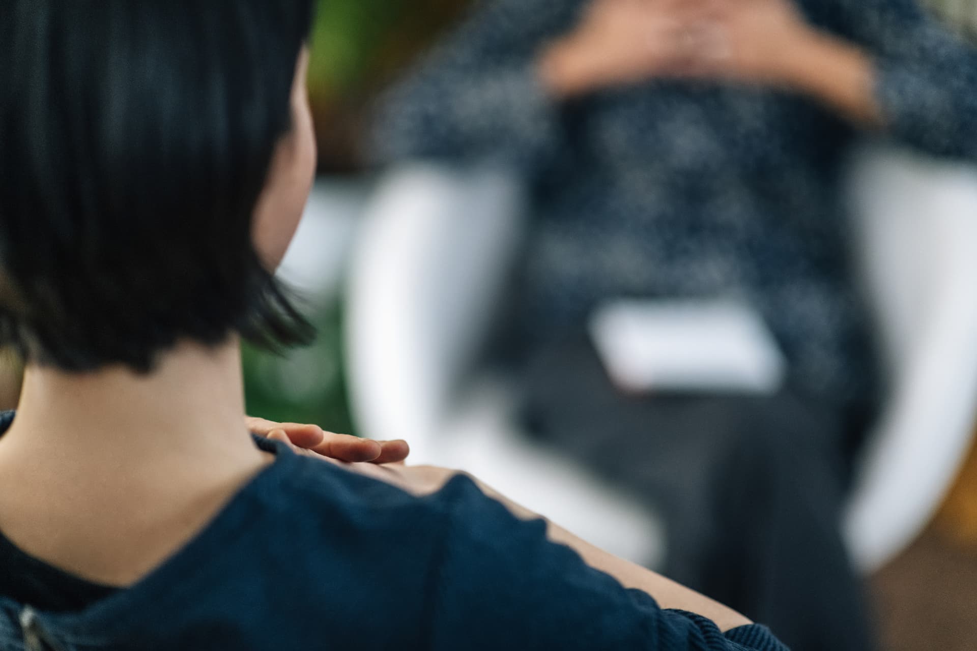 Beratungssituation von hinten: im Vordergrund der dunkle, kinnlange Haarschopf und die Schulter einer Person, auf der eine Hand liegt; unscharf dahinter sitzt eine zweite Person mit gefalteten Händen in einem weißen Sessel.