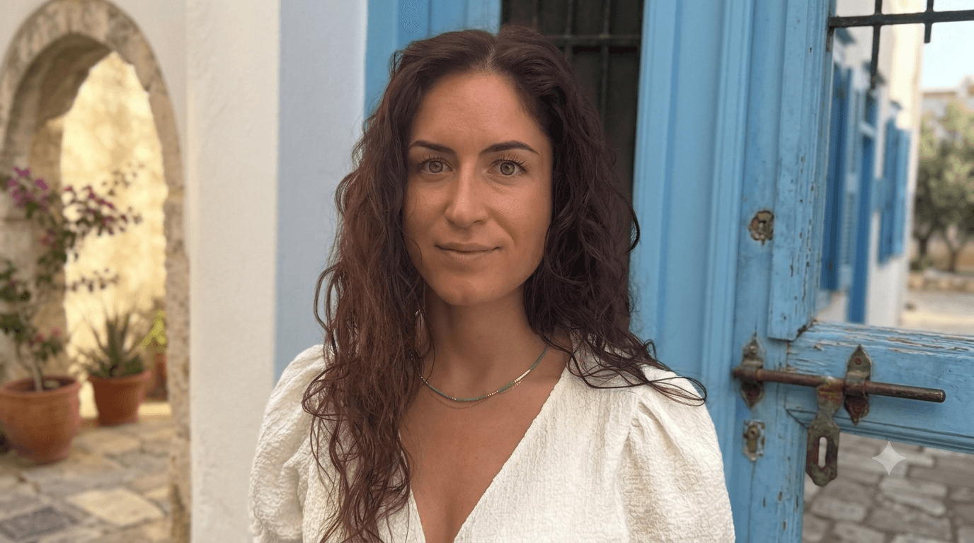 Salome Arnold steht vor einer blau gestrichenen Holztür an einer weiß gekalkten Wand und blickt ruhig in die Kamera. Sie hat lange, dunkelrote Locken und trägt eine cremefarbene Bluse mit feiner Halskette. Warmes Sonnenlicht.