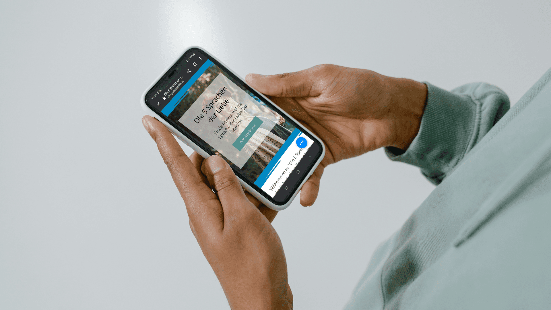 Zwei Hände halten ein Smartphone, auf dem die Startseite des Tests „Die 5 Sprachen der Liebe“ mit einem blauen Knopf zu sehen ist; die Person trägt einen hellgrünen Pullover, der Hintergrund ist hellgrau.