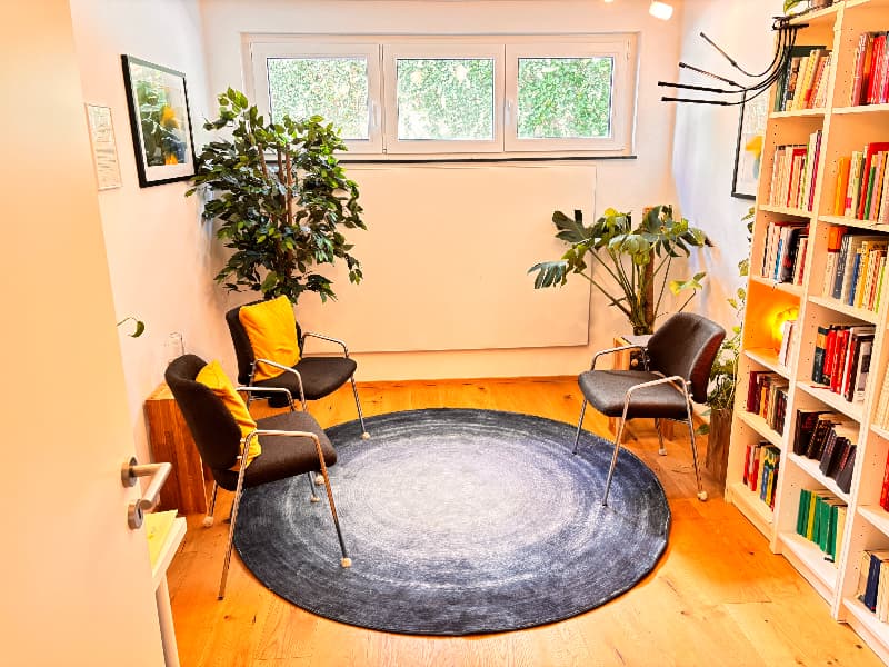 Der Beratungsraum der Praxis: drei dunkle Stühle mit gelben Kissen stehen um einen runden grauen Teppich auf hellem Holzboden. Rechts ein weißes Regal voller Bücher, im Raum ein Ficus und eine Monstera, durch das Fenster fällt Licht auf grüne Blätter.