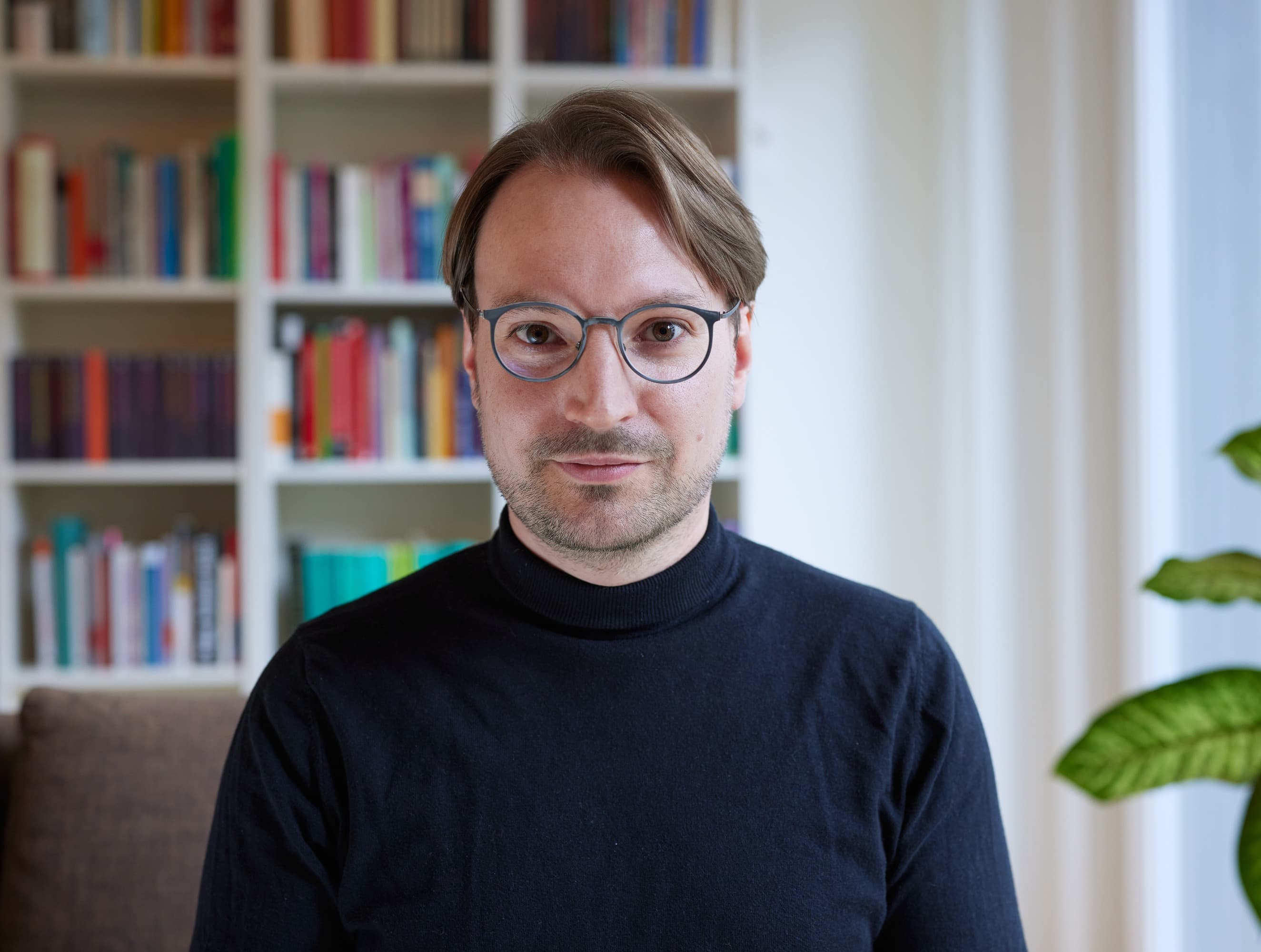 Emanuel Rechsteiner-Grammenos steht vor einem weißen Bücherregal voller bunter Bücher und blickt ruhig in die Kamera. Er trägt eine runde Brille und einen schwarzen Rollkragenpullover; rechts eine Grünpflanze vor einem hellen Fenster.