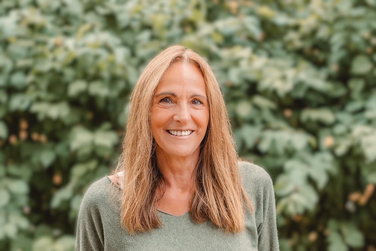 Tatjana Jetter steht vor einer dichten grünen Hecke und lächelt offen in die Kamera. Sie hat lange, hellbraune Haare und trägt einen salbeigrünen Strickpullover. Warmes Tageslicht.