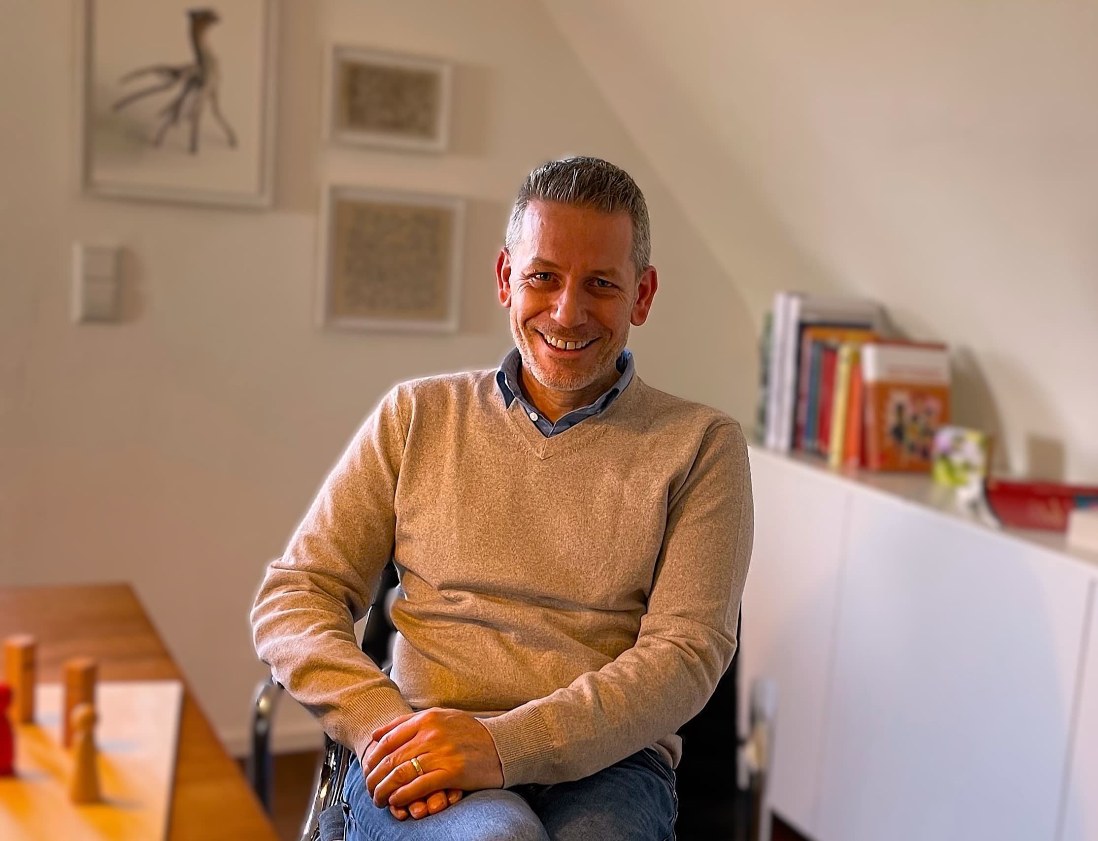 Tobias Jetter sitzt entspannt auf einem Stuhl und lächelt in die Kamera. Er hat kurzes graues Haar und einen kurzen Bart und trägt einen sandfarbenen Pullover über einem blauen Hemd. Hinter ihm ein weißes Sideboard mit bunten Büchern und zwei gerahmte Bilder an der hellen Dachschräge, im Vordergrund helle Holzfiguren auf einem Holztisch.